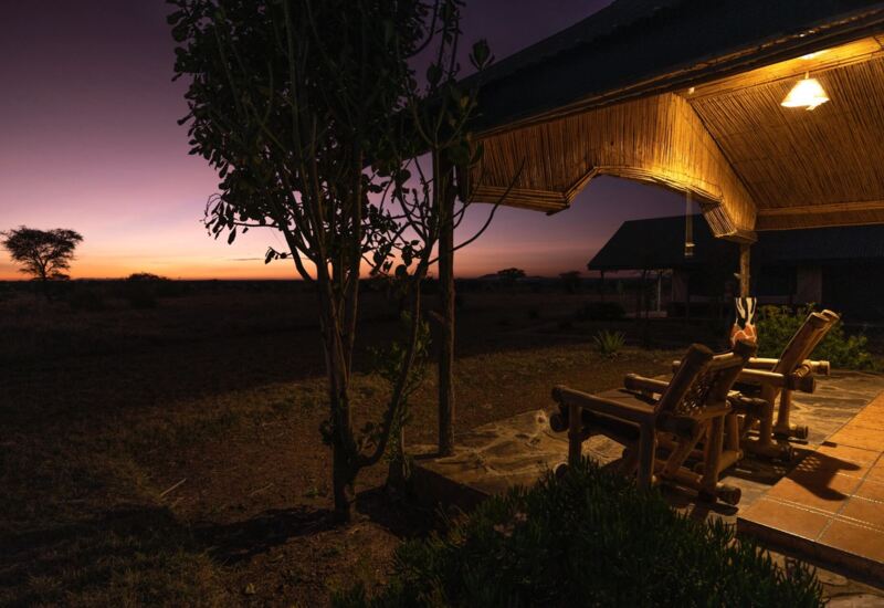 Rustic porch with wooden lounge chairs under warm lighting, overlooking open savannah at sunset at a safari lodge