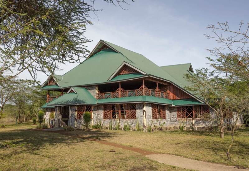 Rustic two-story lodge with green metal roof, stone walls, wooden lattice windows, surrounded by trees and open grassy area