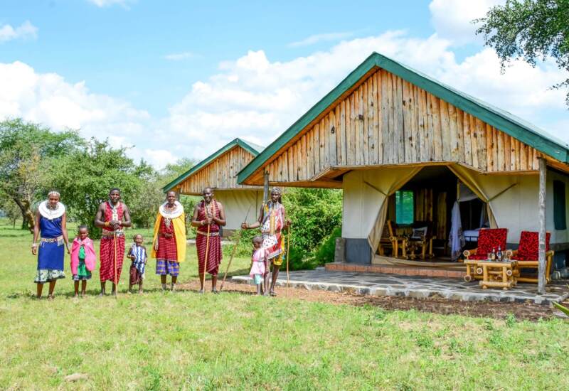 Rustic safari tents with wooden facades and open seating area set in green savannah, with people in traditional Maasai attire nearby