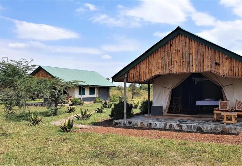 Safari-style tented lodge with wooden porch chairs set on stone platform amid open grassy savanna under blue sky
