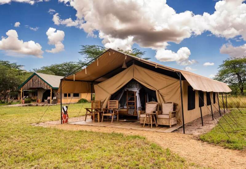 Luxury safari tent with furnished veranda featuring wooden and wicker chairs set in a grassy African landscape under blue sky