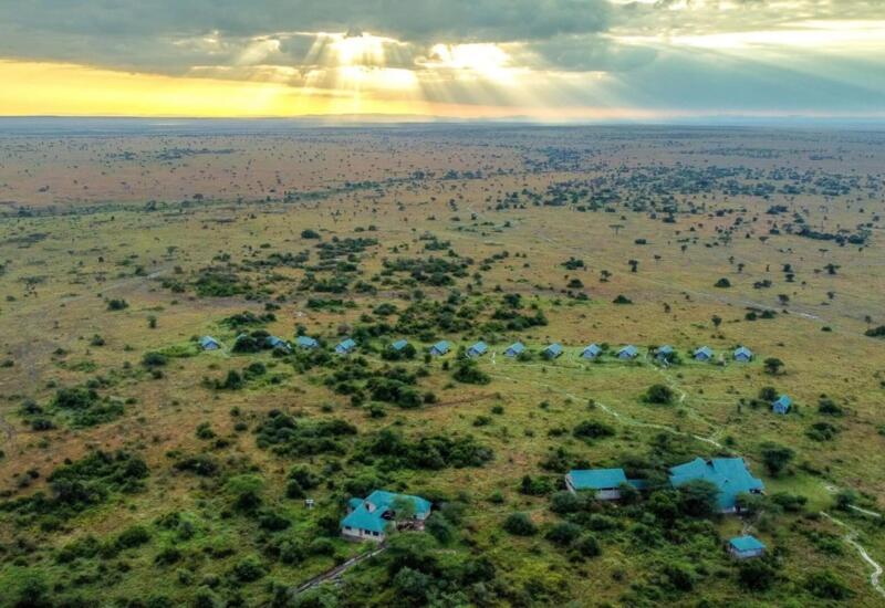 Remote safari lodge with multiple tents and buildings set in vast open savannah under dramatic sun rays breaking through clouds