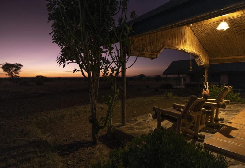 Thatched-roof patio with wooden lounge chairs overlooking African savanna at sunset, illuminated by warm outdoor lighting