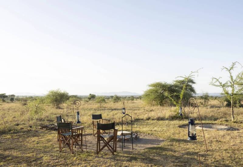 Outdoor safari seating area with wooden chairs around a fire pit, lanterns hanging on metal stands, set in open savannah landscape