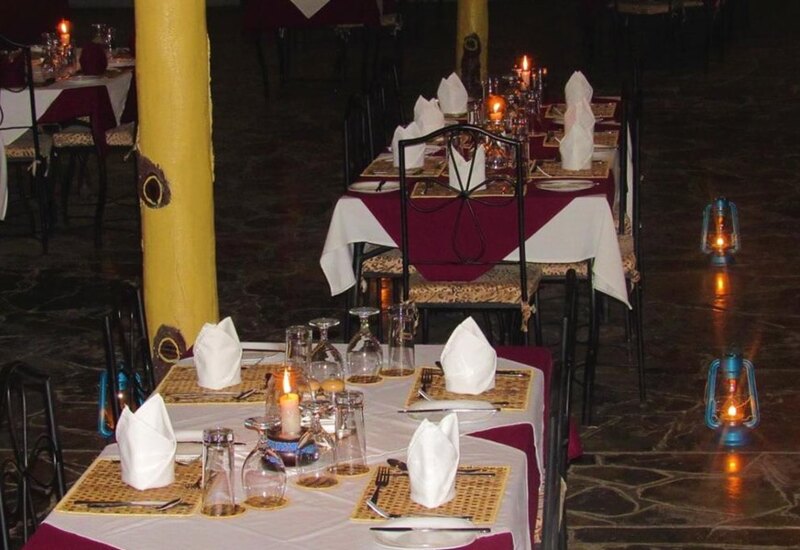 Cozy indoor dining area with tables set in white and burgundy linens, candles, bottles, and folded napkins in rustic ambiance