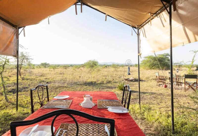 Outdoor dining area under canvas canopy with red tablecloth and views of open savannah landscape and distant hills