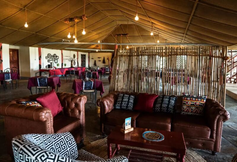 Cozy lounge area with leather sofas, patterned cushions, and a rustic dining room featuring tables with maroon tablecloths and eclectic decor