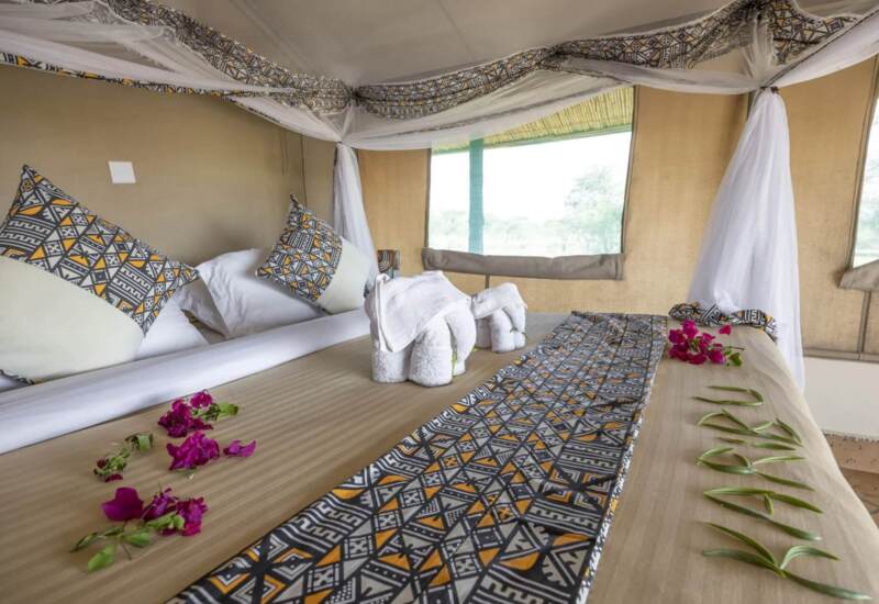 Safari tent bedroom with patterned pillows, mosquito netting, towel elephants, and fresh flowers on beige bedding