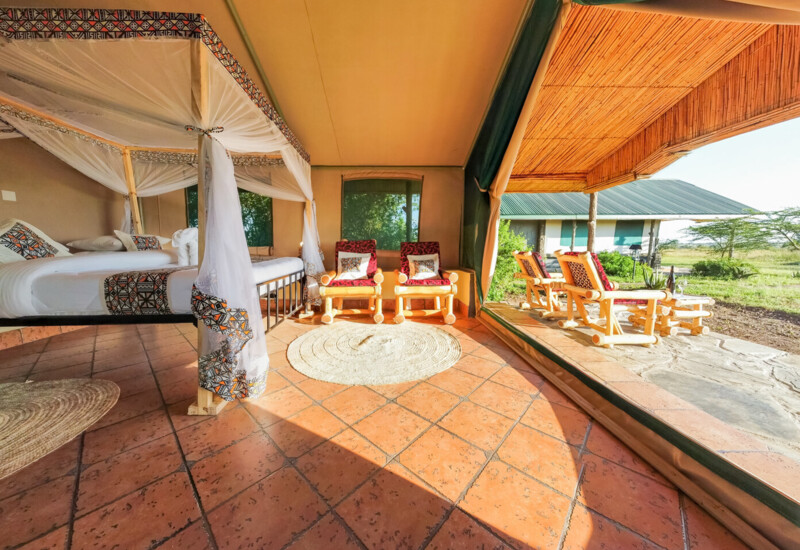 Spacious safari tent with canopy bed, patterned linens, wooden chairs inside and outside on stone patio, overlooks grassy savanna