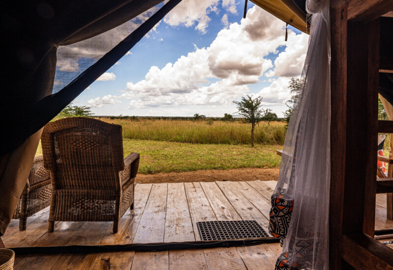 Wooden deck with wicker chair overlooking open grassland under a partly cloudy sky from a luxury safari tent