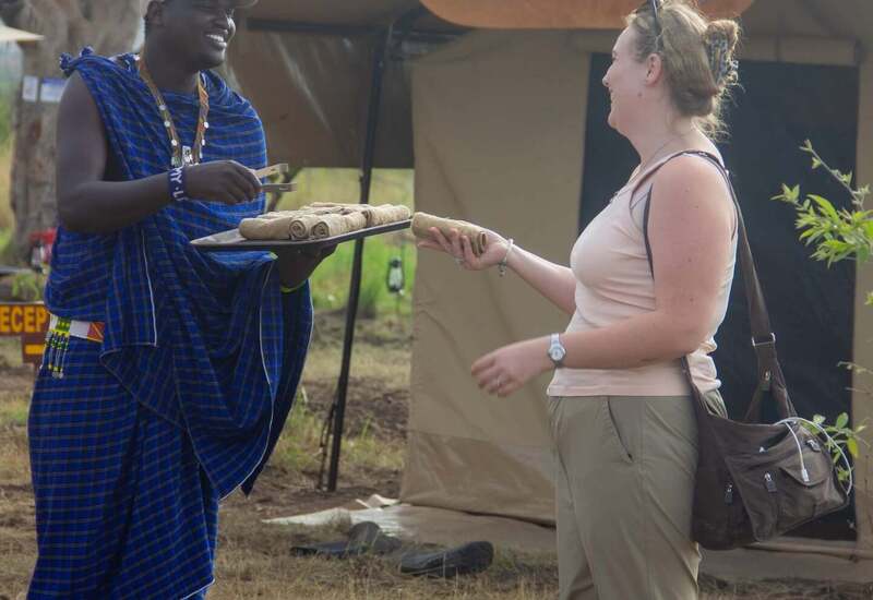 Maasai guide offers rolled welcome towels to smiling guest at tented Serengeti safari camp entrance.
