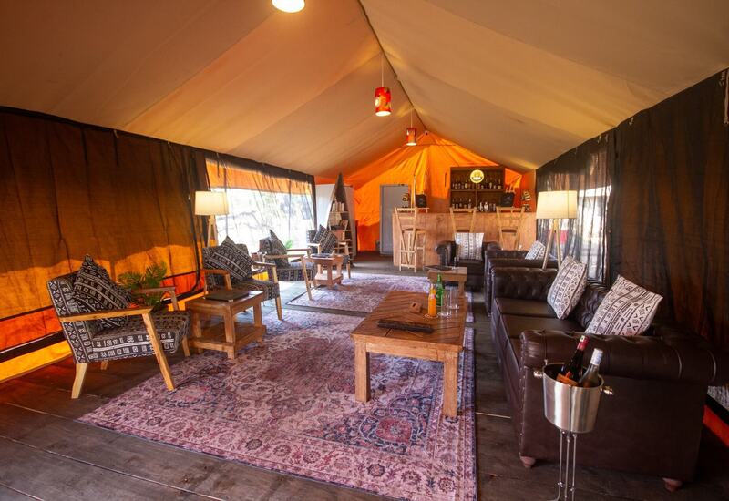 Spacious luxury safari tent lounge with leather sofas, patterned armchairs, wooden coffee tables, bar counter, rugs and warm pendant lighting.