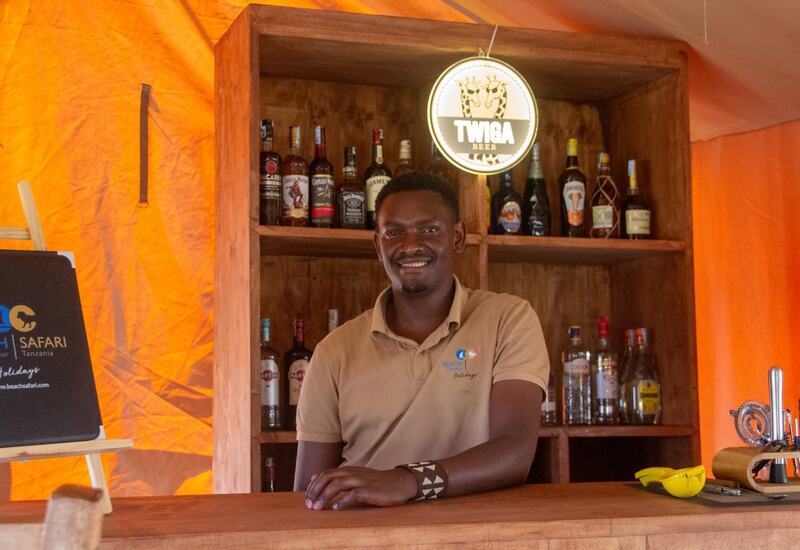 Smiling bartender in beige polo behind a wooden hotel bar with spirits, Twiga beer sign and warm orange tent