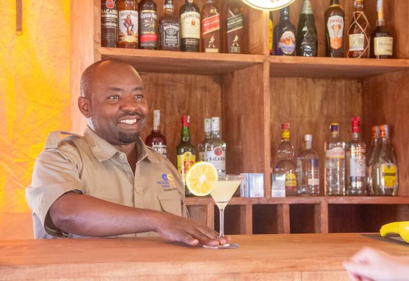 Smiling bartender at a hotel beach bar serving a lemon-garnished cocktail, wooden shelves of spirits behind him.