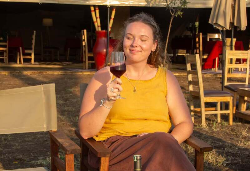 Guest relaxing on outdoor lodge terrace at golden hour, sipping red wine in a wooden chair beside a bottle, canvas dining tent behind.