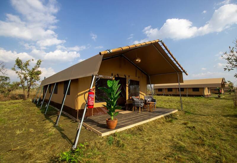 Tented safari cabin with shaded wooden deck, potted plant and two chairs for outdoor lounging under a blue sky.