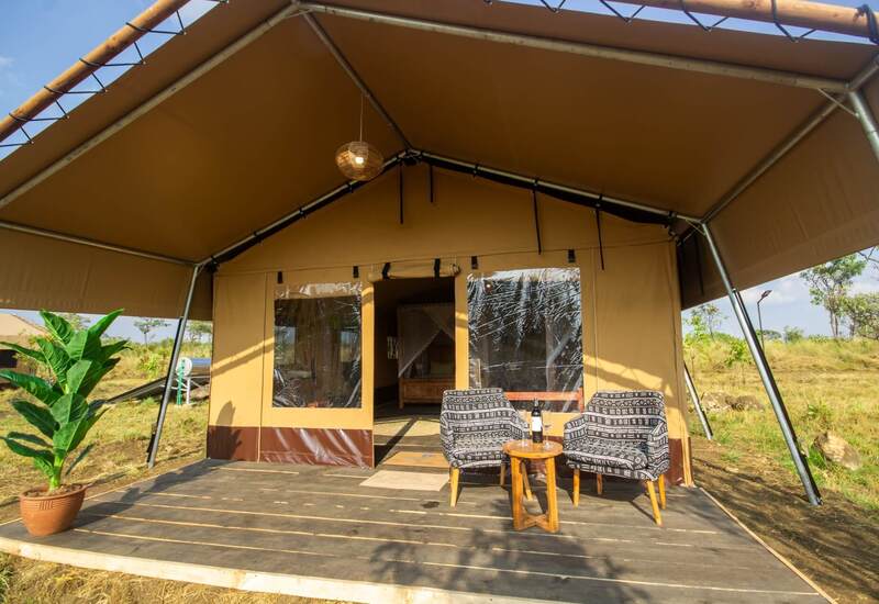 Luxury canvas safari tent with shaded porch, two patterned lounge chairs and table with wine, hanging rattan lamp and grassy views.