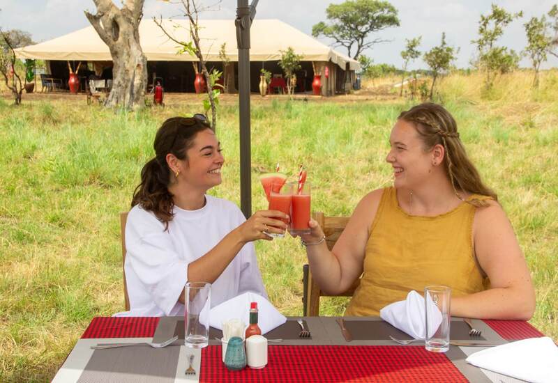 Two women toasting watermelon cocktails at an outdoor dining table in front of a canvas safari tent on grassy savanna, table set with napkins and condiments.