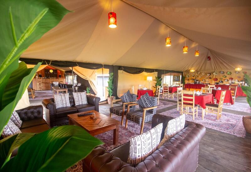 Luxury safari tent lounge and dining space with leather sofas, patterned armchairs, wooden tables, red-tablecloth dining, buffet, pendant lights and potted plants.