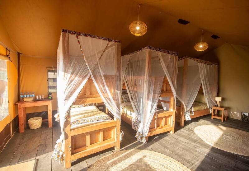 Shared safari-style tent with wooden four-poster beds draped in white mosquito nets, woven pendant lights, bedside tables and bottled water.