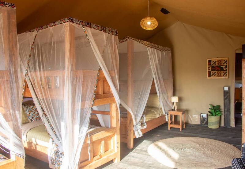 Safari-style tent bedroom with wooden bunk beds draped in white mosquito nets, patterned cushions, bedside lamp, woven pendant and round jute rug.