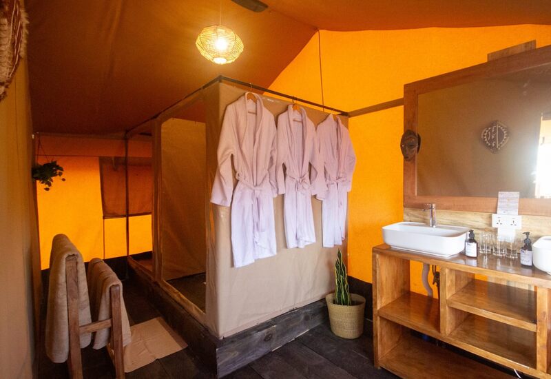 Warm glamping tent bathroom with hanging white robes, wooden vanity and basin sink, towel rack, pendant light, potted plant
