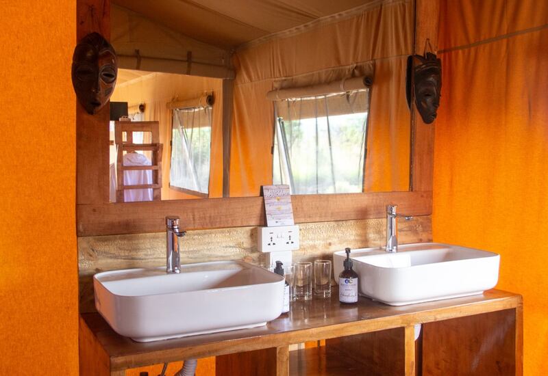 Double white sinks on a wooden vanity in an orange canvas safari tent, mirror reflecting bunks and carved masks.