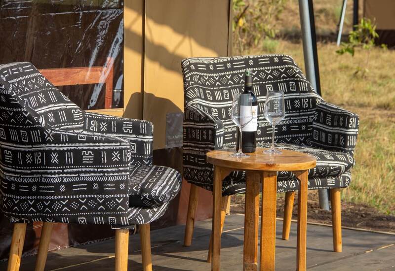 Two patterned lounge chairs on a tented glamping deck with a wooden side table holding a bottle of wine and two glasses
