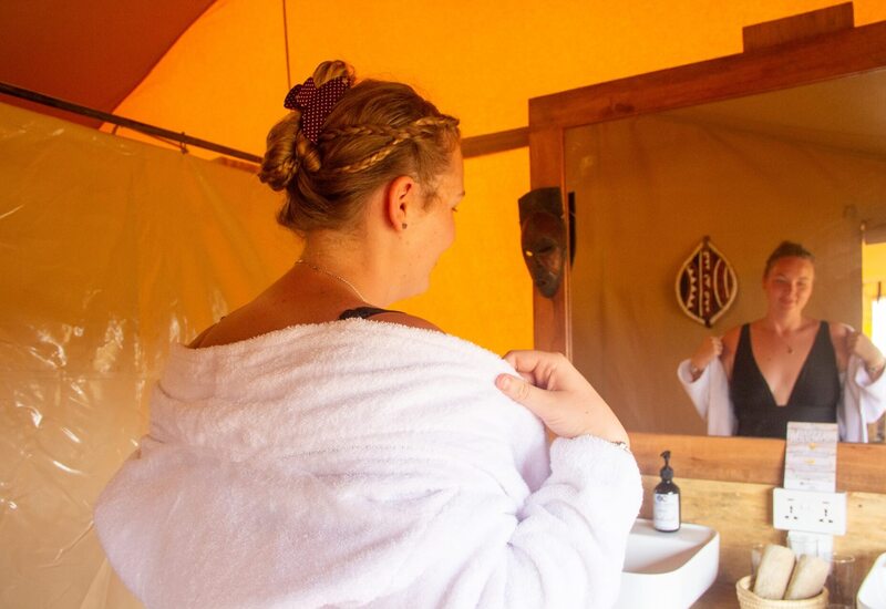 Guest in plush robe adjusting towel in warm-lit glamping bathroom with wooden mirror, sink, soap and woven towel basket