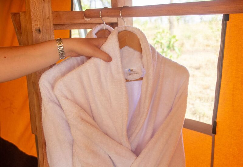 Plush white bathrobe on wooden hanger in a warm tented room, hand adjusting it — cozy spa amenity