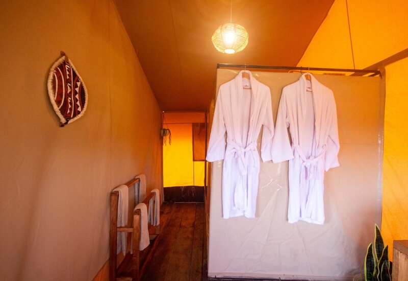 Glamping tent interior with two white bathrobes hanging on a divider, warm rattan pendant light, wooden floor and towel racks