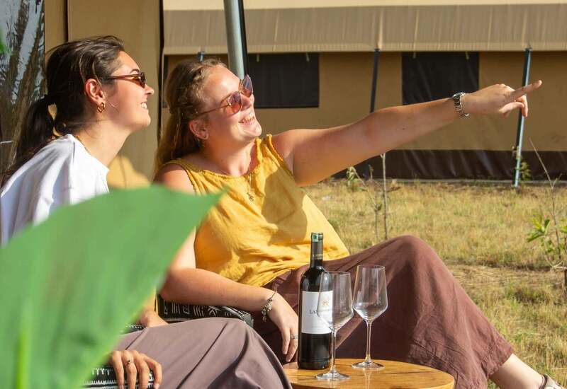 Two women in a shaded glamping lounge smiling and pointing, wine bottle and glasses on a small wooden table, relaxed outdoor vibe