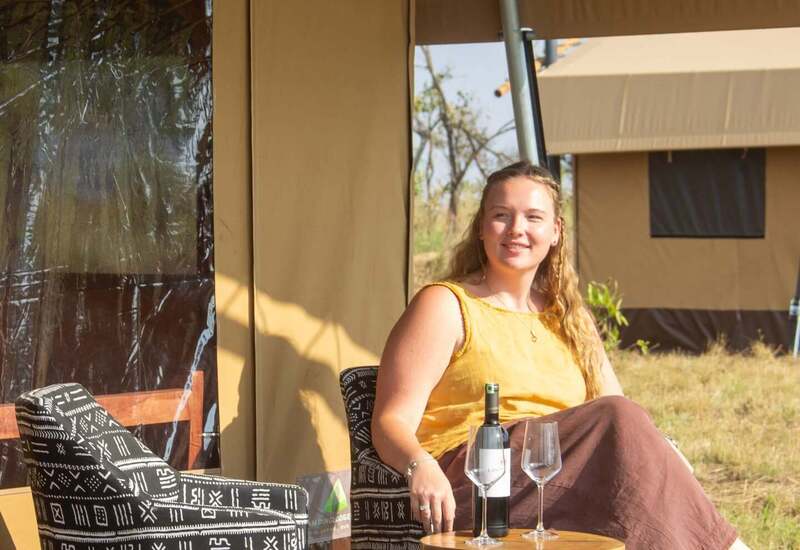 Sunlit guest relaxing on a safari tent deck in patterned armchair, wine bottle and two glasses on small wooden table, canvas tents nearby.