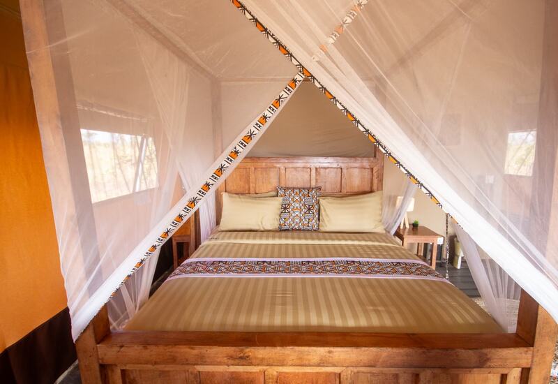 Four-poster wooden bed in a safari tent draped with white mosquito net, striped linens, patterned throw and bedside lamp.