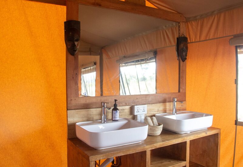 Safari tent bathroom with twin white vessel sinks on wood vanity, large mirror, woven towels and orange canvas walls.