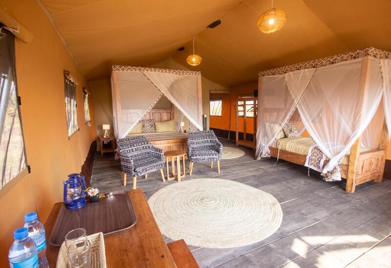 Spacious luxury safari tent with two mosquito-net four-poster beds, wooden furnishings, patterned armchairs, woven rugs and warm pendant lighting.