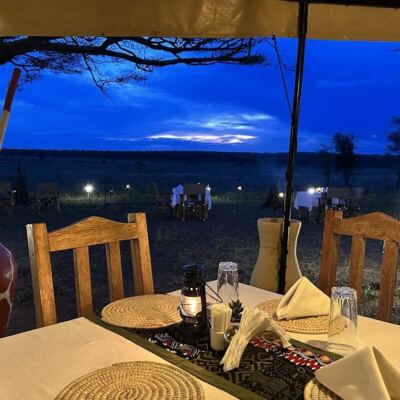 Outdoor dining setup with wooden chairs, woven placemats, lantern, and sunset views over open savannah landscape
