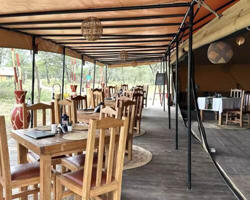 Rustic open-air dining area with wooden tables and chairs under a canopy, surrounded by natural savannah landscape