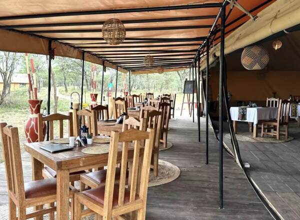 Rustic open-air dining area with wooden tables and chairs under a canopy, surrounded by natural savannah landscape