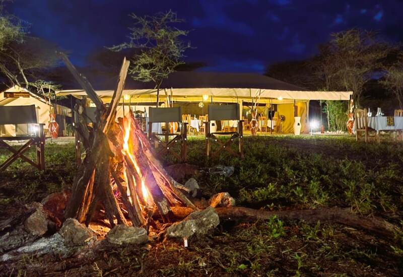 Outdoor night scene at safari lodge with campfire, canvas dining tent, wooden chairs, and soft ambient lighting