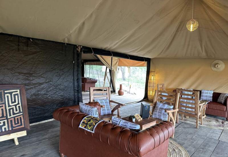 Cozy safari tent lounge with leather sofas, patterned cushions, wooden chairs, warm lighting, and rustic decor on wooden flooring