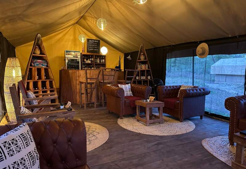 Cozy glamping lounge with leather armchairs, wooden bar, woven rugs, ambient lighting, and large screened window overlooking nature.