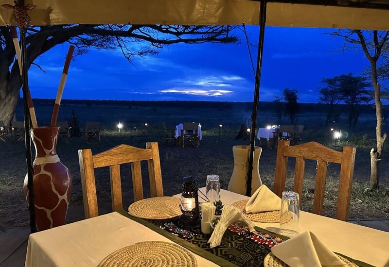 Outdoor dining setup with wooden chairs, woven placemats, lantern, and sunset views over open savannah landscape