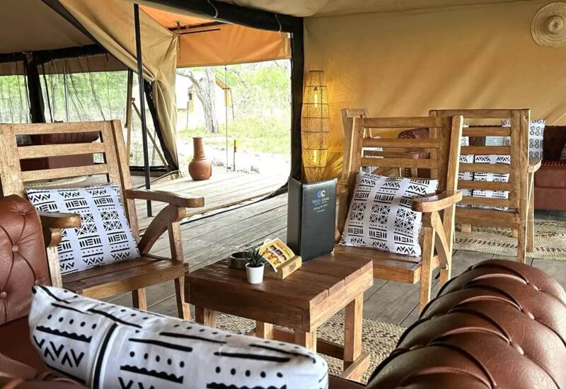 Outdoor lounge area with wooden chairs, patterned cushions, leather sofas, woven lamp, and rustic wooden deck in a tented setting