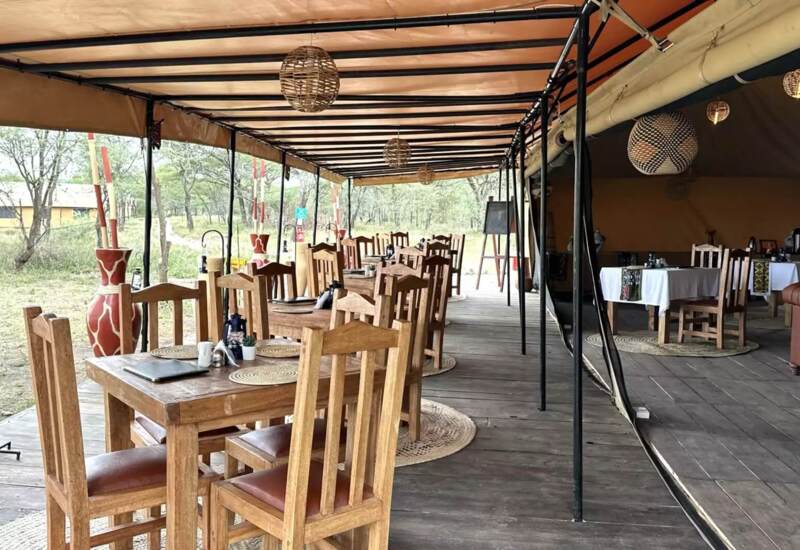 Rustic open-air dining area with wooden tables and chairs under a canopy, surrounded by natural savannah landscape