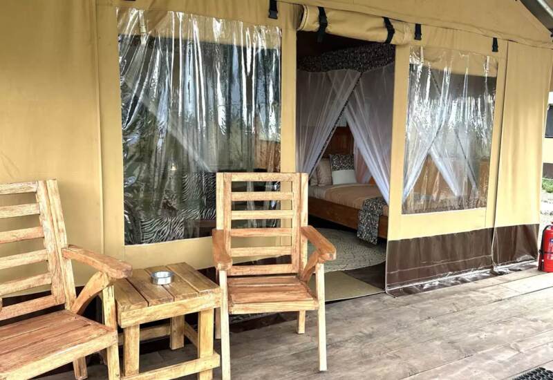 Outdoor wooden chairs and table on tent porch with clear vinyl windows revealing a bed with mosquito netting inside