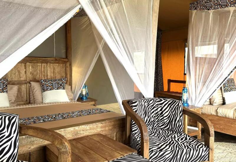 Rustic hotel tent room with wooden furniture, double bed with patterned pillows, mosquito nets, zebra-print chairs, and soft natural light