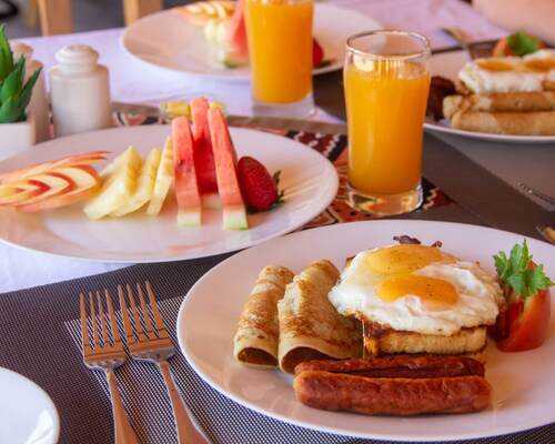 Hearty hotel breakfast with fried eggs on toast, sausages and rolled crepes, fresh fruit slices and glasses of orange juice
