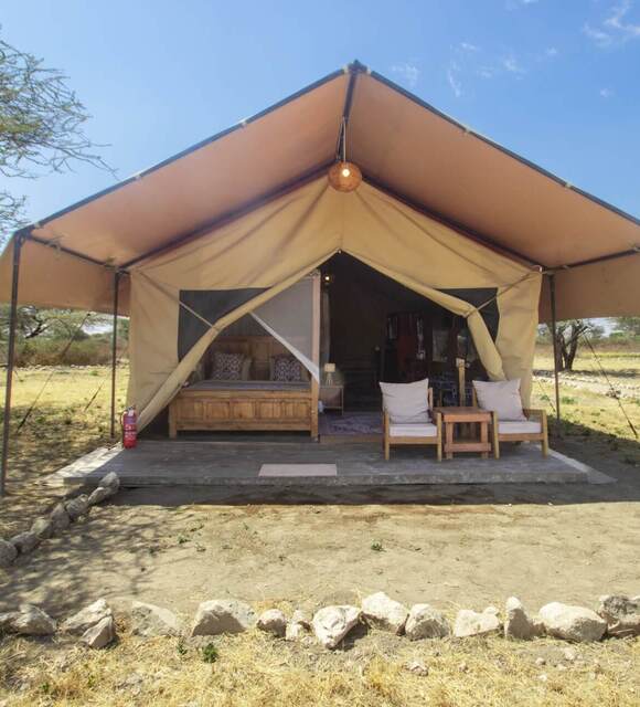 Canvas safari tent with shaded wooden porch, king bed with mosquito net, two cushioned chairs, lantern and dry savannah view.