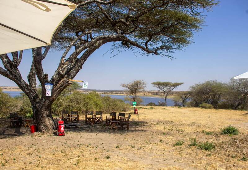 Shaded outdoor lounge with wooden director chairs and umbrellas beneath an acacia canopy, overlooking a tranquil river.
